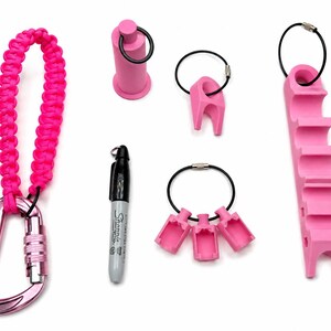 May include: A set of pink accessories including a paracord lanyard with a carabiner, a cylindrical holder, a single-slot holder, a triple-slot holder, and a long multi-slot holder, all attached to black wire rings, alongside a black Sharpie marker.