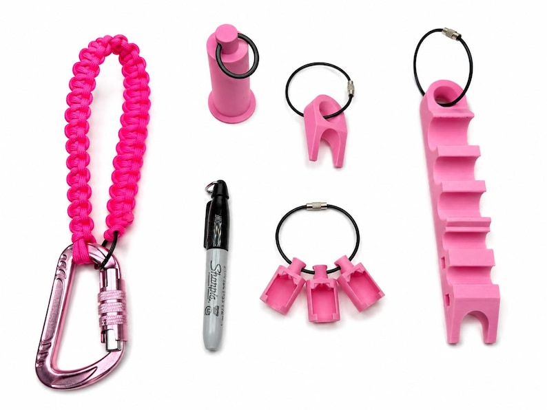 May include: A set of pink accessories including a paracord lanyard with a carabiner, a cylindrical holder, a single-slot holder, a triple-slot holder, and a long multi-slot holder, all attached to black wire rings, alongside a black Sharpie marker.