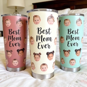 May include: Three stainless steel tumblers with clear lids, each featuring a repeating pattern of a child's face and the black script text Best Mom Ever. The tumblers are colored in muted rose, cream, and light mint green with silver bases.