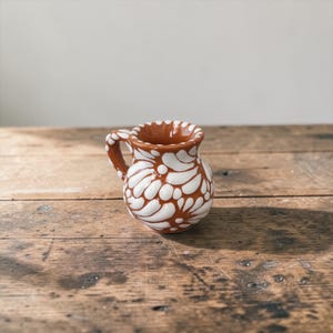 May include: A small, brown and white ceramic pitcher with a handle. The pitcher is decorated with a white floral pattern. The pitcher sits on a rustic wooden surface.
