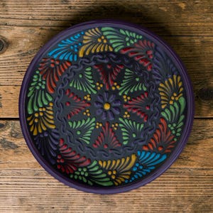 May include: A round, dark purple ceramic plate with a raised, colorful floral design. The design features a central flower and radiating patterns in red, yellow, green, and blue. The plate sits on a wooden surface.
