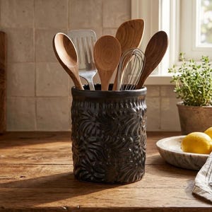 May include: A black ceramic utensil crock with a textured, embossed leaf pattern on the exterior. The cylindrical container holds a collection of wooden spoons, a metal slotted turner, and a metal wire whisk, all standing upright inside.