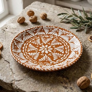 May include: A decorative ceramic plate with a brown base and white floral pattern. The plate features a detailed design of stylized flowers and leaves. The plate is round and appears to be handmade. Walnuts and greenery are in the background.