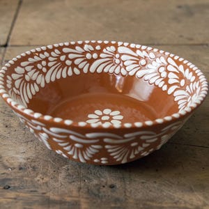 May include: A terracotta-colored ceramic bowl with a white floral pattern around the rim and a central flower design. The bowl has a glossy finish and sits on a wooden surface. The decorative bowl is suitable for serving food.