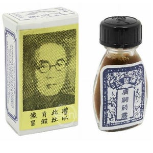 May include: A small glass bottle with a black cap and a label with Asian characters, next to a rectangular box with a portrait and text. The bottle contains a brown liquid. The box has text in English and Asian characters.