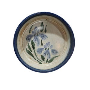 May include: A round ceramic bowl with a cream-colored interior featuring a hand-painted blue and green floral design. The bowl has a thick, dark blue rim and a smooth, glazed finish, showcasing a simple, functional design with visible brushstrokes.