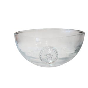 Peut inclure: Un bol en verre transparent avec un bord lisse et arrondi et un médaillon circulaire en relief sur le côté extérieur. Le médaillon présente un motif texturé rappelant une fleur, avec un groupe central de petits points entouré de lignes radiales.