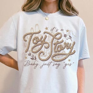 May include: A light blue t-shirt featuring a graphic design with the text "it's a Toy Story" in a rope-like font, accompanied by desert-themed illustrations of cacti and mountains, with the additional text "Baby, just say yes" below.