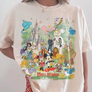 May include: Ivory-colored t-shirt featuring a colorful graphic of Disney characters and the text "Magic Kingdom". The design includes Mickey and Minnie Mouse, Aladdin, and other beloved characters. The shirt is labeled "Comfort Colors".