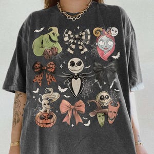 May include: A charcoal gray oversized t-shirt featuring a graphic print of characters from The Nightmare Before Christmas, including Jack Skellington, Sally, Zero, and Oogie Boogie, interspersed with various bows, spiderwebs, and bats.