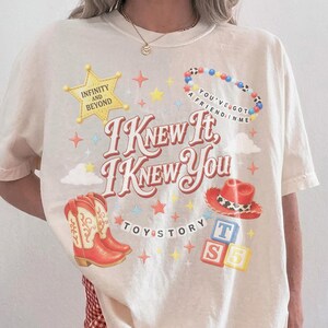 May include: An ivory short-sleeve t-shirt featuring a graphic print with the text "I KNEW IT, I KNEW YOU", "INFINITY AND BEYOND", "YOU'VE GOT A FRIEND IN ME", "TOY STORY", and "T S". The design includes a yellow star, red cowboy boots, a red hat, and blocks.