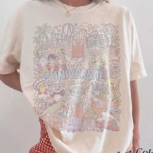 K&ouml;nnte beinhalten: Elfenbeinfarbenes T-Shirt mit einer farbenfrohen Grafik von Universal Studios-Charakteren und -Attraktionen. Das Shirt tr&auml;gt den Text "UNIVERSAL Studios" in einer Pastellschrift. Das Shirt ist von der Marke Comfort Colors.