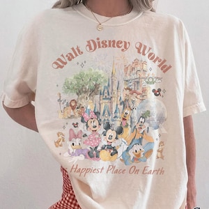 May include: Ivory-colored t-shirt with a Walt Disney World graphic featuring various cartoon characters and the text "Happiest Place On Earth." The shirt is a Comfort Colors brand.