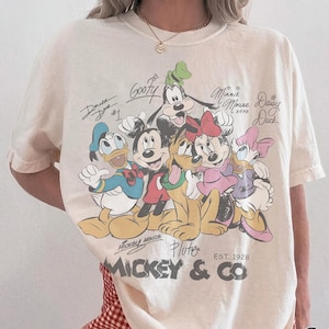 K&ouml;nnte beinhalten: Elfenbeinfarbenes T-Shirt mit einer Grafik von Mickey Mouse und seinen Freunden, darunter Donald Duck, Goofy und Minnie Mouse. Das Shirt hat den Text "Mickey & Co" und "EST. 1928". Das Shirt ist mit "Comfort Colors" und "Ivory" gekennzeichnet.