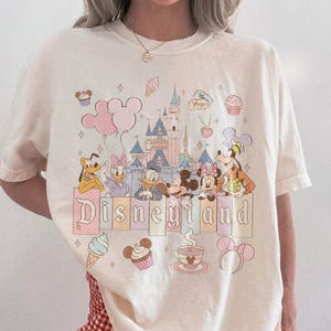 Może przedstawiać: Kremowa koszulka z grafiką Disneyland z postaciami z kresk&oacute;wek i zamkiem. Projekt zawiera słowo "Disneyland" w pastelowej czcionce, a także babeczki, lody i balony. Koszulka jest oznaczona jako "Comfort Colors Ivory".