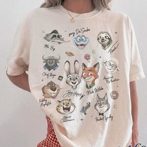 May include: An ivory short-sleeve t-shirt featuring a graphic print of various animated animal characters including a rabbit, fox, sloth, and sheep. The text includes names like Judy Hopps, Nick Wilde, Mr. Big, and Flash, accompanied by small star icons.