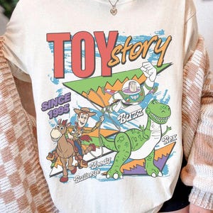 May include: Cream-colored t-shirt featuring a graphic design from the movie "Toy Story." The design includes the text "TOY Story" in red and yellow, along with images of Woody, Buzz Lightyear, Rex, and Bullseye. The text "SINCE 1995" is also present.
