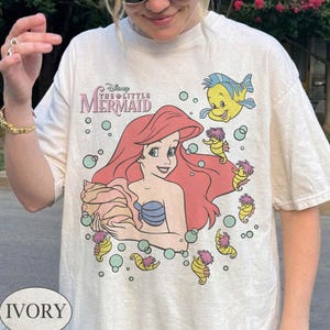 May include: Ivory t-shirt with a graphic of Ariel from Disney's The Little Mermaid. The design includes Ariel with red hair, a blue seashell bra, and a seashell. The shirt also features Flounder and seahorses.