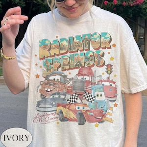 K&ouml;nnte beinhalten: Elfenbeinfarbenes T-Shirt mit einem farbenfrohen Aufdruck von Zeichentrickfiguren und dem Text "Radiator Springs" und "Lightning McQueen". Das Design umfasst verschiedene Zeichentrickautos, Sterne und eine Ampel.