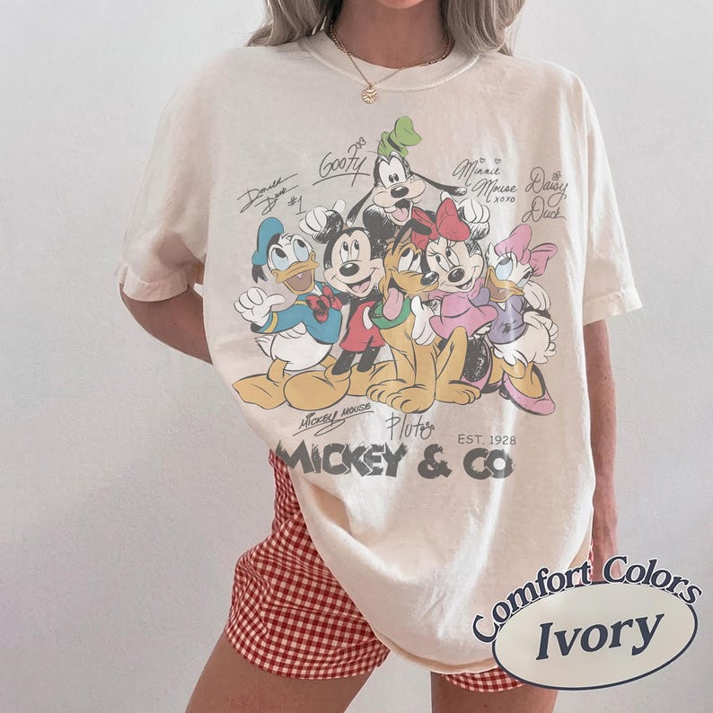 K&ouml;nnte beinhalten: Elfenbeinfarbenes T-Shirt mit einer Grafik von Mickey Mouse und seinen Freunden, darunter Donald Duck, Goofy und Minnie Mouse. Das Shirt hat den Text "Mickey & Co" und "EST. 1928". Das Shirt ist mit "Comfort Colors" und "Ivory" gekennzeichnet.