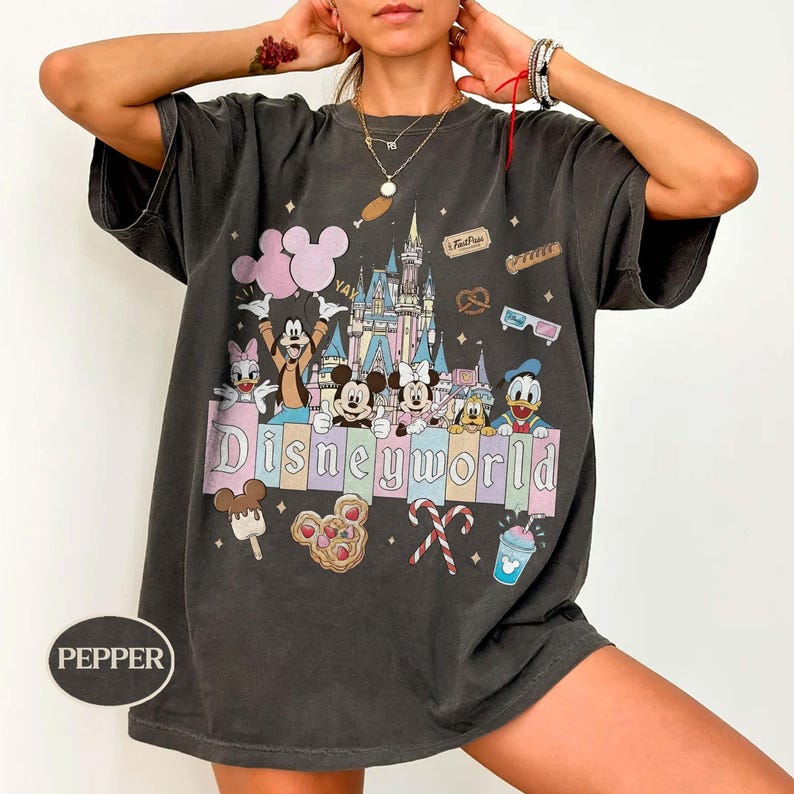 May include: Dark grey t-shirt with a Disney World graphic. The design features Mickey and Minnie Mouse, Donald Duck, Goofy, and Daisy Duck, a castle, balloons, and food icons. The word "Disneyworld" is displayed in a colourful font.