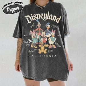 May include: Dark grey oversized t-shirt with a Disneyland graphic featuring Disney characters, the Disneyland logo, and the text "California". The shirt includes the text "EST. 1955" and the Comfort Colors Pepper logo.