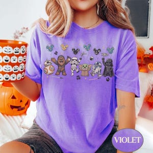 May include: A violet t-shirt featuring a graphic of Star Wars characters holding balloons. The characters include BB-8, Chewbacca, a Stormtrooper, Grogu, R2-D2, and Darth Vader. The word "VIOLET" is printed on the bottom right.
