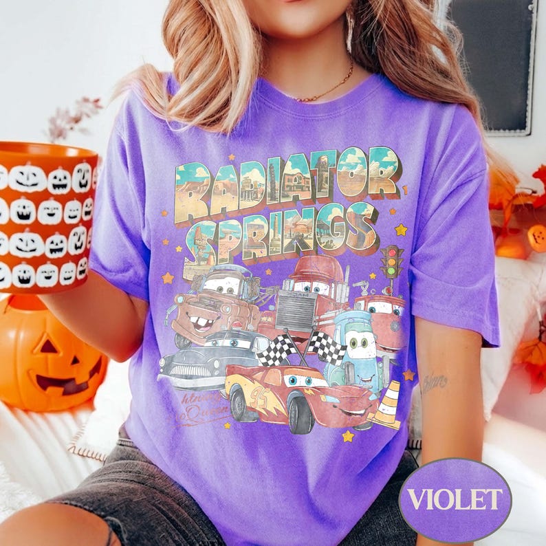 K&ouml;nnte beinhalten: Violettes T-Shirt mit einem Aufdruck von Charakteren aus dem Film Cars und dem Text "Radiator Springs". Das Design umfasst Autocharaktere, eine Ampel und Zielflaggen. Das T-Shirt hat einen Rundhalsausschnitt und kurze &Auml;rmel.