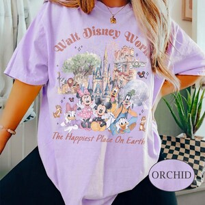 May include: Orchid-colored t-shirt featuring a graphic of Disney characters and the text "Walt Disney World" and "The Happiest Place On Earth." The design includes Mickey Mouse, Minnie Mouse, Donald Duck, and other characters.