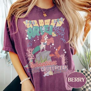 May include: A berry-colored short-sleeve t-shirt featuring a graphic print of Ariel, Flounder, and Sebastian. The text reads Atlantica Ariel Ocean Tour 1991, Sebastian and the Hot Crustacean Band. The design includes coral, bubbles, and a rock.