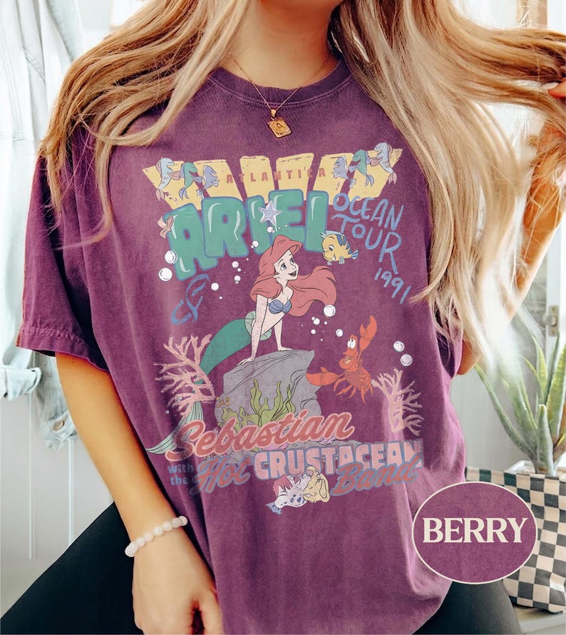 May include: A berry-colored short-sleeve t-shirt featuring a graphic print of Ariel, Flounder, and Sebastian. The text reads Atlantica Ariel Ocean Tour 1991, Sebastian and the Hot Crustacean Band. The design includes coral, bubbles, and a rock.