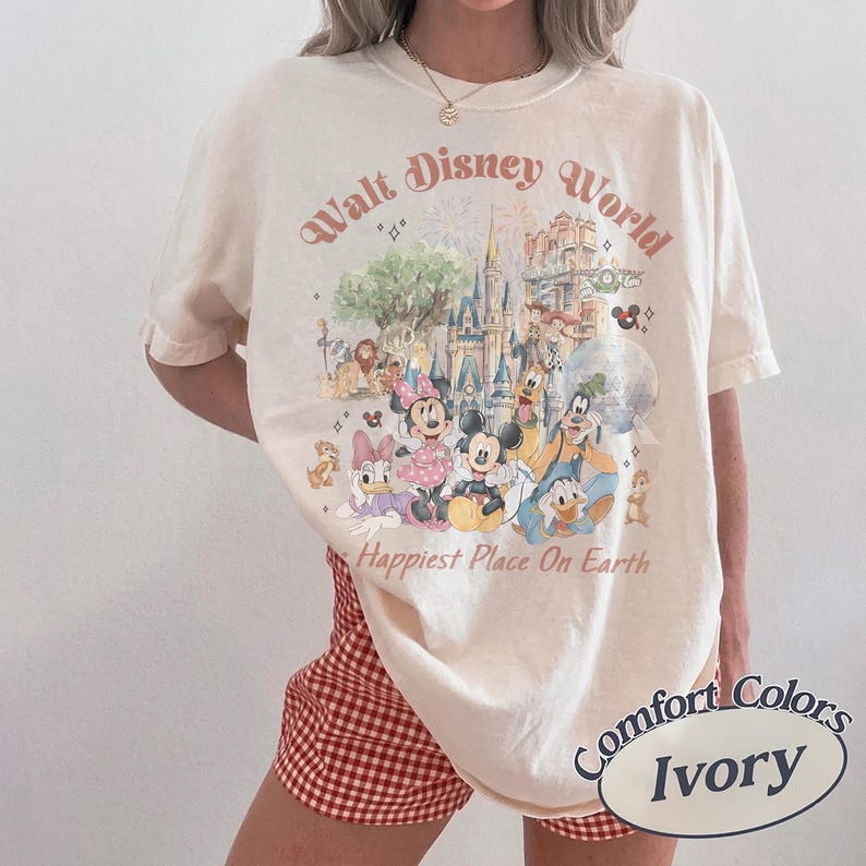 May include: Ivory-colored t-shirt with a Walt Disney World graphic featuring various cartoon characters and the text "Happiest Place On Earth." The shirt is a Comfort Colors brand.