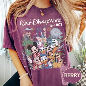 May include: A berry-colored t-shirt featuring a graphic print of Walt Disney World Est. 1971 with illustrations of Mickey Mouse, Minnie Mouse, Donald Duck, Daisy Duck, Goofy, Pluto, Chip, and Dale taking a selfie in front of park landmarks.
