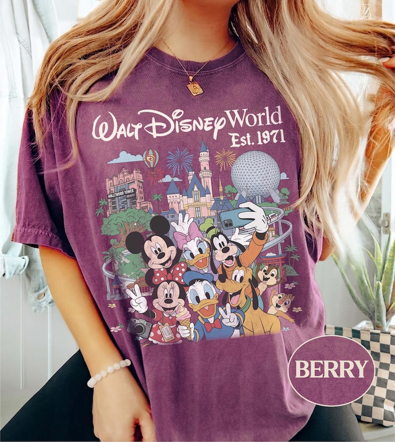 May include: A berry-colored t-shirt featuring a graphic print of Walt Disney World Est. 1971 with illustrations of Mickey Mouse, Minnie Mouse, Donald Duck, Daisy Duck, Goofy, Pluto, Chip, and Dale taking a selfie in front of park landmarks.