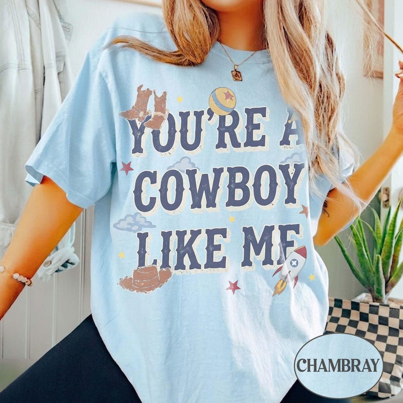 May include: A light blue t-shirt featuring the dark blue text "YOU'RE A COWBOY LIKE ME" surrounded by illustrations of cowboy boots, a hat, a rocket, a ball, stars, and clouds. The shirt has a crew neckline and short sleeves.