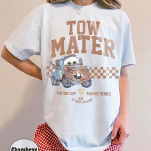 May include: A light blue t-shirt featuring the text TOW MATER in brown letters above a graphic of the character Tow Mater. Below the character is a checkered pattern, the text PISTON CUP RACING SERIES, a trophy icon, and 5 TIME CHAMP.