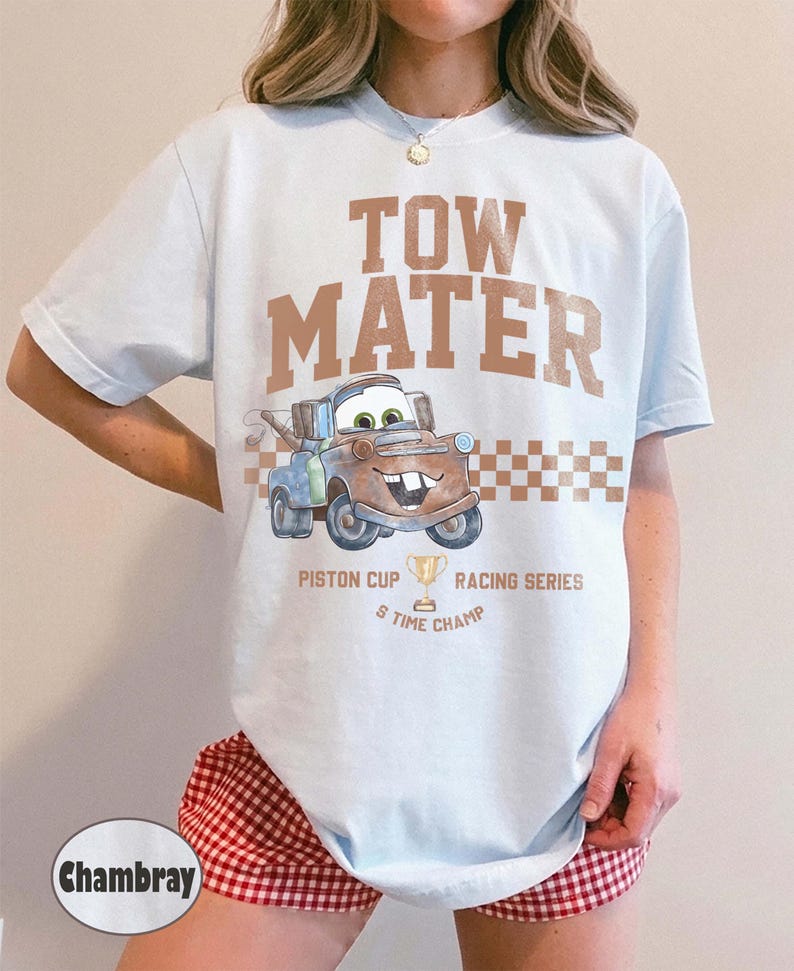 May include: A light blue t-shirt featuring the text TOW MATER in brown letters above a graphic of the character Tow Mater. Below the character is a checkered pattern, the text PISTON CUP RACING SERIES, a trophy icon, and 5 TIME CHAMP.