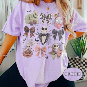 May include: A light purple t-shirt featuring a graphic print of characters and bows from The Nightmare Before Christmas, including Jack Skellington, Sally, Oogie Boogie, and Zero. The design includes spiderwebs, bats, and the word ORCHID.
