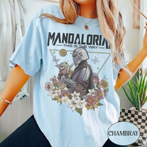 Op de afbeelding: Lichtblauw T-shirt met de tekst "MANDALORIA - THIS IS THE WAY." Het ontwerp toont een Mandalorian karakter en Grogu, omringd door bloemen en vlinders. Het shirt is chambray van kleur.