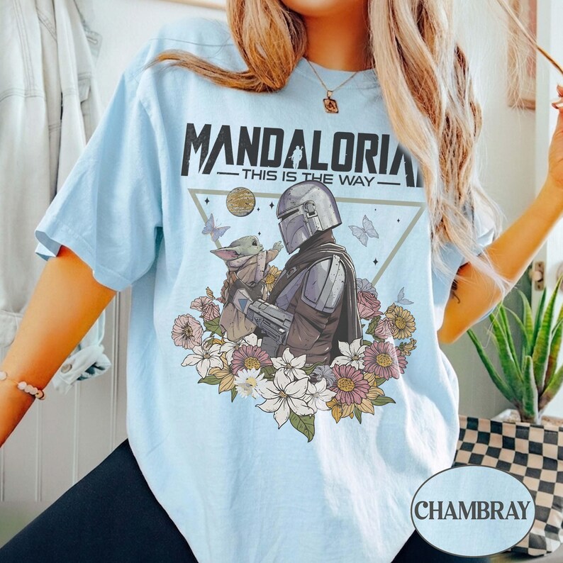 Op de afbeelding: Lichtblauw T-shirt met de tekst "MANDALORIA - THIS IS THE WAY." Het ontwerp toont een Mandalorian karakter en Grogu, omringd door bloemen en vlinders. Het shirt is chambray van kleur.