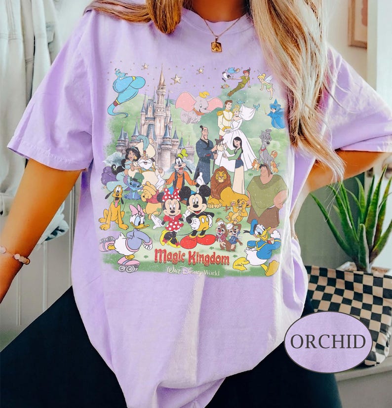May include: A light purple t-shirt featuring a colorful graphic of Disney characters and the text "Magic Kingdom". The design includes Mickey and Minnie Mouse, Donald Duck, and other beloved characters. The shirt is labeled "Orchid".