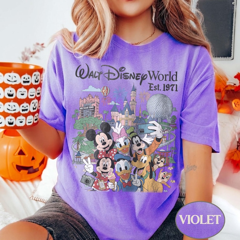 May include: A violet t-shirt featuring a graphic print of Walt Disney World Est. 1971 with Mickey Mouse, Minnie Mouse, Donald Duck, Daisy Duck, Goofy, Pluto, Chip, and Dale in front of park landmarks including the castle and Epcot sphere.