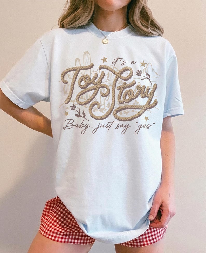 May include: A light blue t-shirt featuring a graphic design with the text "it's a Toy Story" in a rope-like font, accompanied by desert-themed illustrations of cacti and mountains, with the additional text "Baby, just say yes" below.