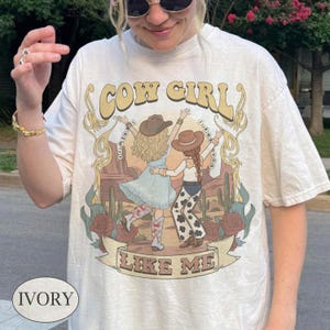 Puede incluir: Una camiseta de manga corta color marfil con un estampado gr&aacute;fico de dos figuras con sombreros de vaquero y ropa del oeste bailando en un paisaje des&eacute;rtico con cactus y rosas. El texto dice COW GIRL LIKE ME en una fuente estilizada.