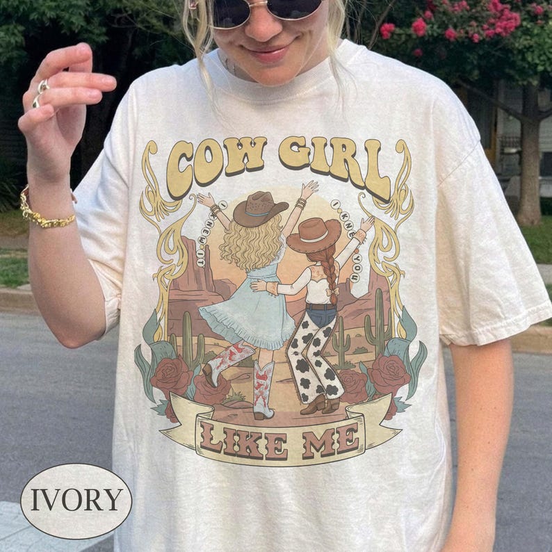 Puede incluir: Una camiseta de manga corta color marfil con un estampado gr&aacute;fico de dos figuras con sombreros de vaquero y ropa del oeste bailando en un paisaje des&eacute;rtico con cactus y rosas. El texto dice COW GIRL LIKE ME en una fuente estilizada.