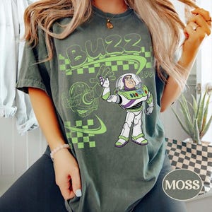 May include: A dark green t-shirt featuring a graphic of the character Buzz Lightyear in his white, green, and purple space suit. The design includes the word BUZZ in large green letters, a green checkered pattern, and a faint outline of the character.