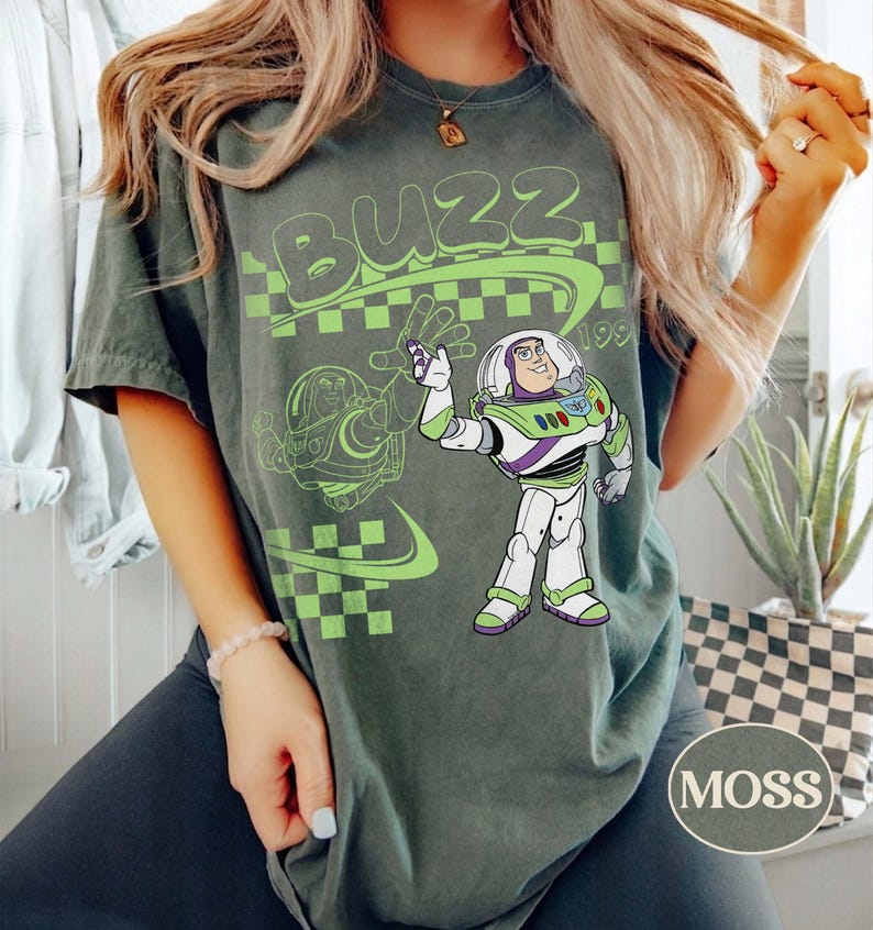 May include: A dark green t-shirt featuring a graphic of the character Buzz Lightyear in his white, green, and purple space suit. The design includes the word BUZZ in large green letters, a green checkered pattern, and a faint outline of the character.