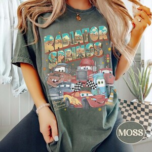 K&ouml;nnte beinhalten: Dunkelgr&uuml;nes T-Shirt mit dem Schriftzug "Radiator Springs" und einer Grafik von animierten Autos. Das Design umfasst Charaktere wie Mater und Lightning McQueen. Das Shirt hat einen Vintage-Look.