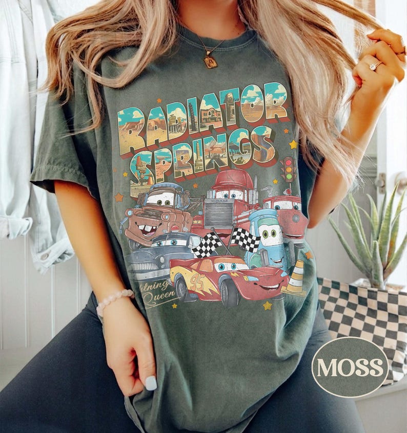 K&ouml;nnte beinhalten: Dunkelgr&uuml;nes T-Shirt mit dem Schriftzug "Radiator Springs" und einer Grafik von animierten Autos. Das Design umfasst Charaktere wie Mater und Lightning McQueen. Das Shirt hat einen Vintage-Look.