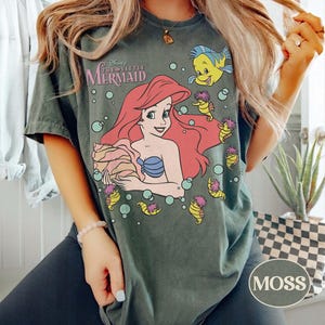 May include: Moss green t-shirt with a graphic of Ariel from Disney's The Little Mermaid. The design includes Ariel with red hair, a blue seashell bra, and Flounder. The shirt has the text "Disney The Little Mermaid".